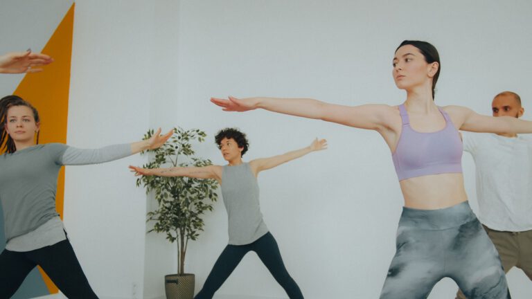 Vier Personen in Sportkleidung stehen in einem hellen Raum in einer Yoga-Haltung mit seitlich ausgestreckten Armen